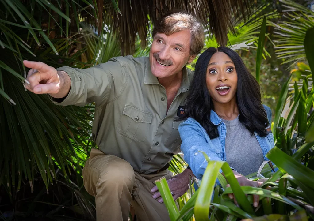 A man in a khaki shirt points excitedly while kneeling in tropical foliage beside a woman with long dark hair, who looks surprised and delighted. Both are surrounded by green leaves.